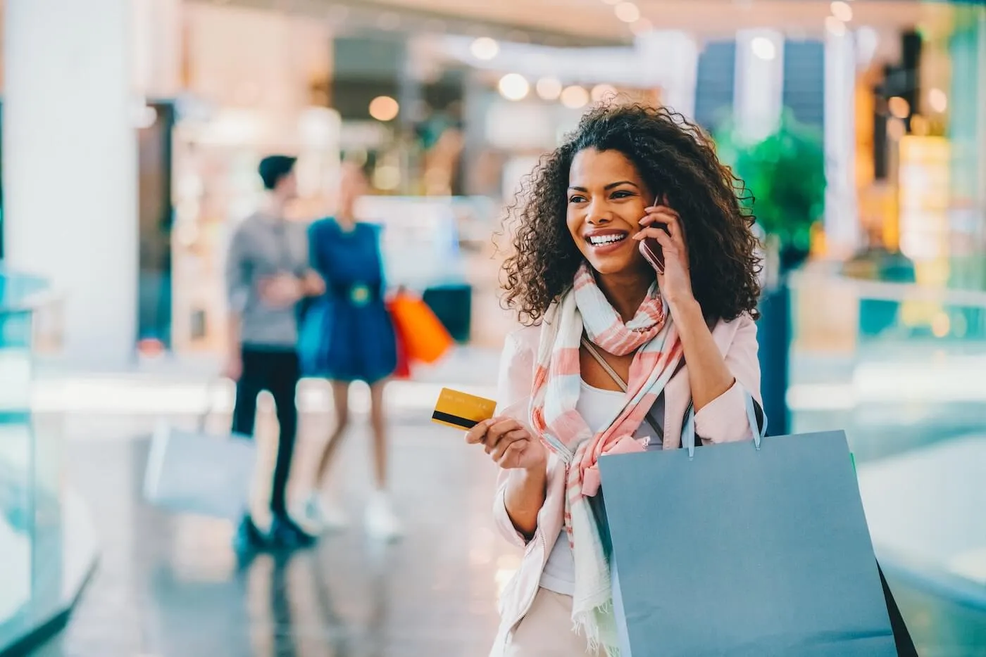 Happy young woman with a shopping bag holding a credit card and making a phone call in the mall