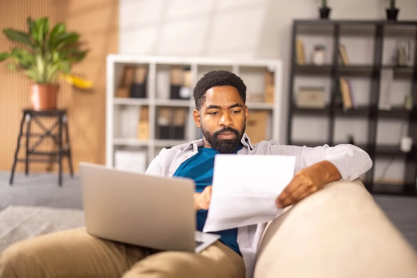 Focused man lying on a sofa with a laptop, reviewing the printed documents