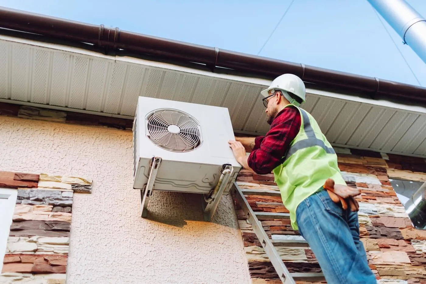 A handyman in the uniform standing on a ladder and fixing an outdoor AC unit.