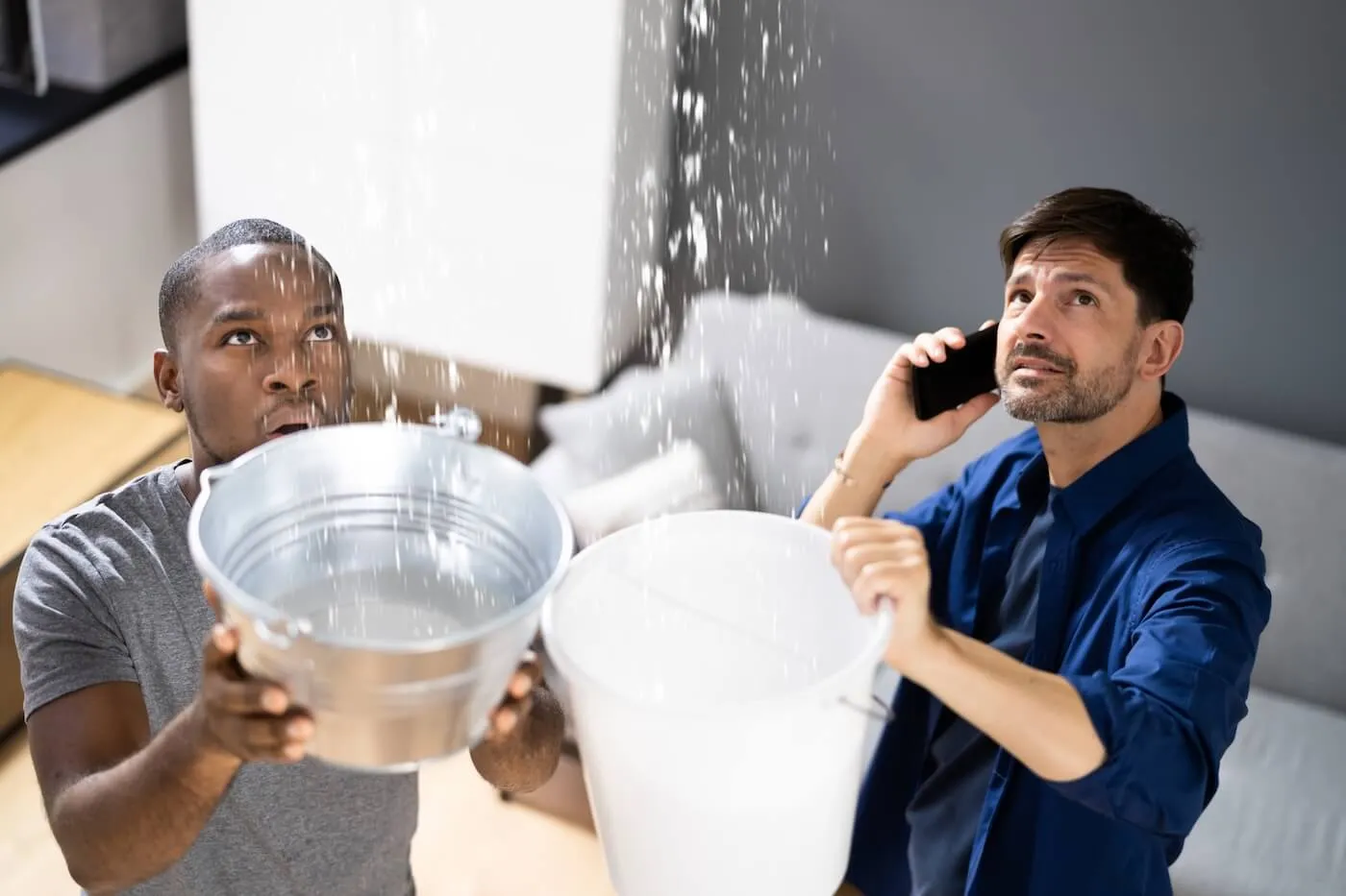 Two frustrated young men catch water from a ceiling leak in containers; one is calling insurance company