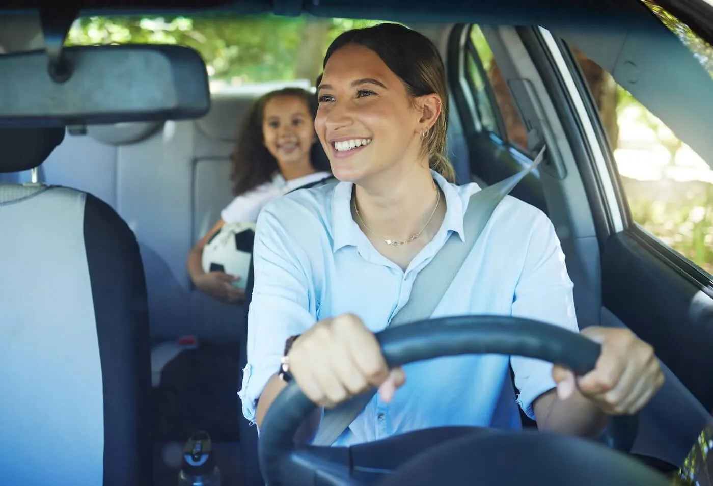 A woman smiles in a ear view mirror to a young girl with a soccer ball on a back seat while driving a car