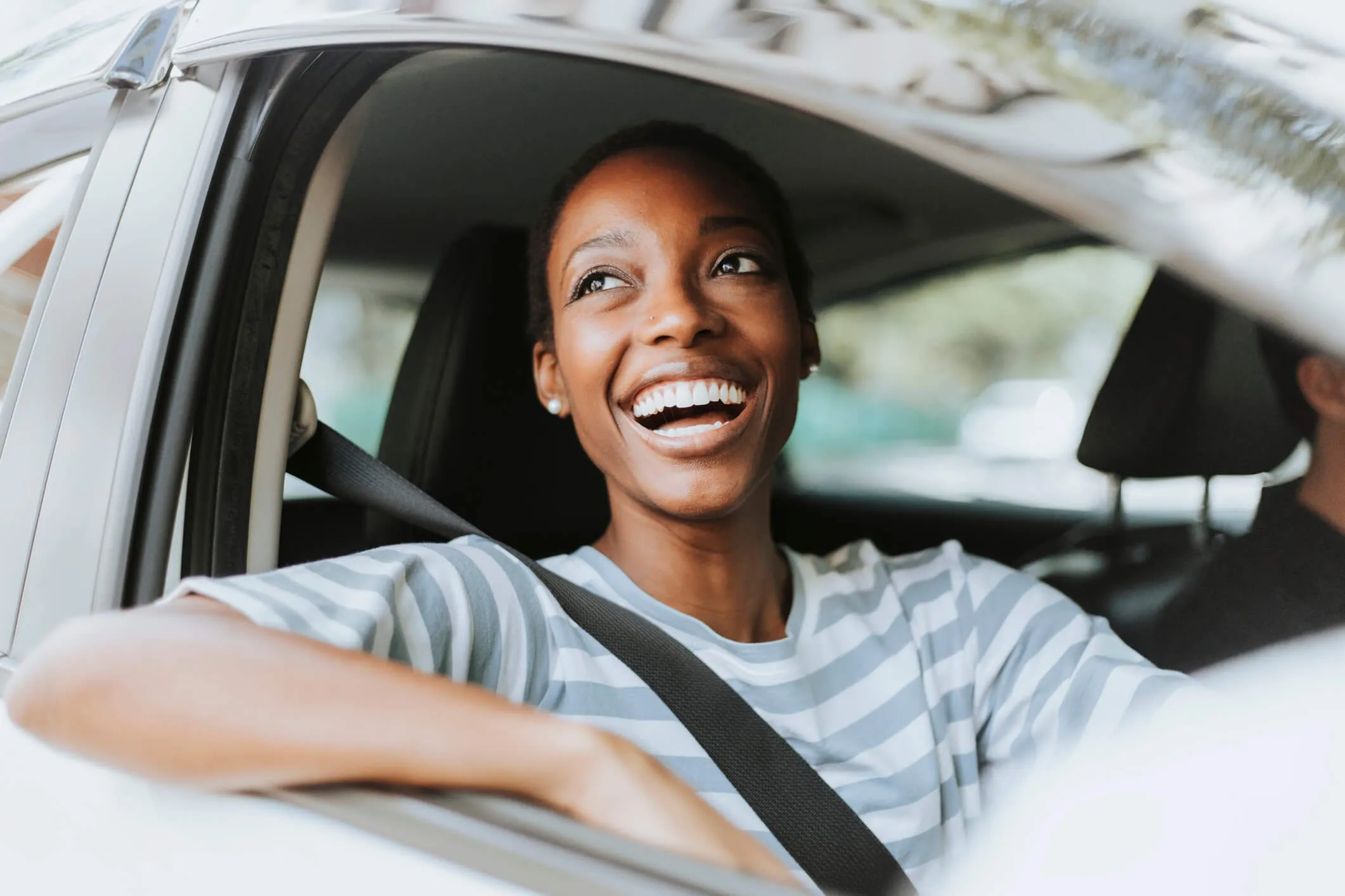 A smiling woman driving a car on a nice day with the window rolled down.