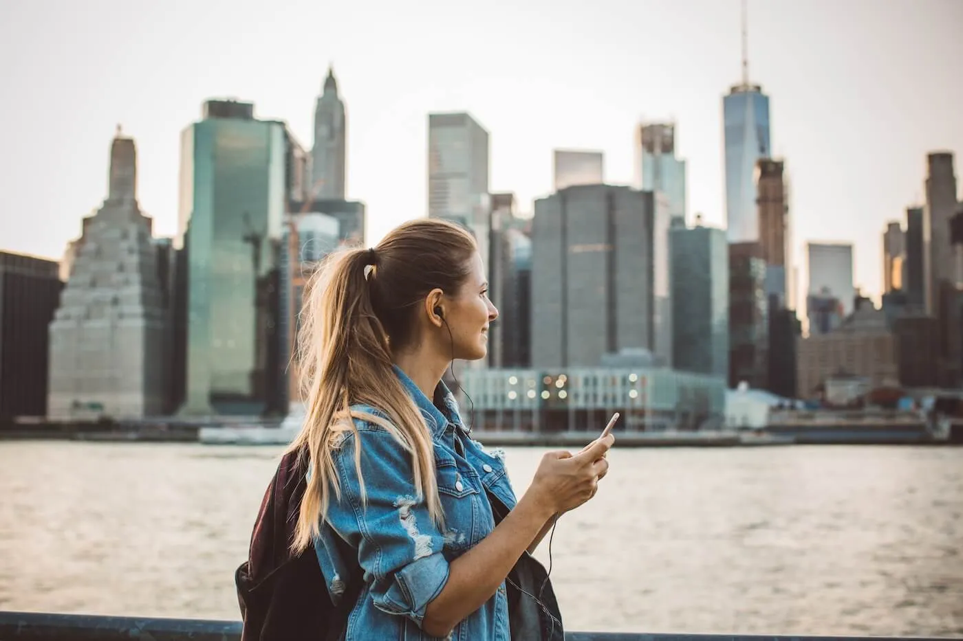 Young woman wearing a backpack enjoying the scenic view of New York City across the water while holding a smartphone.