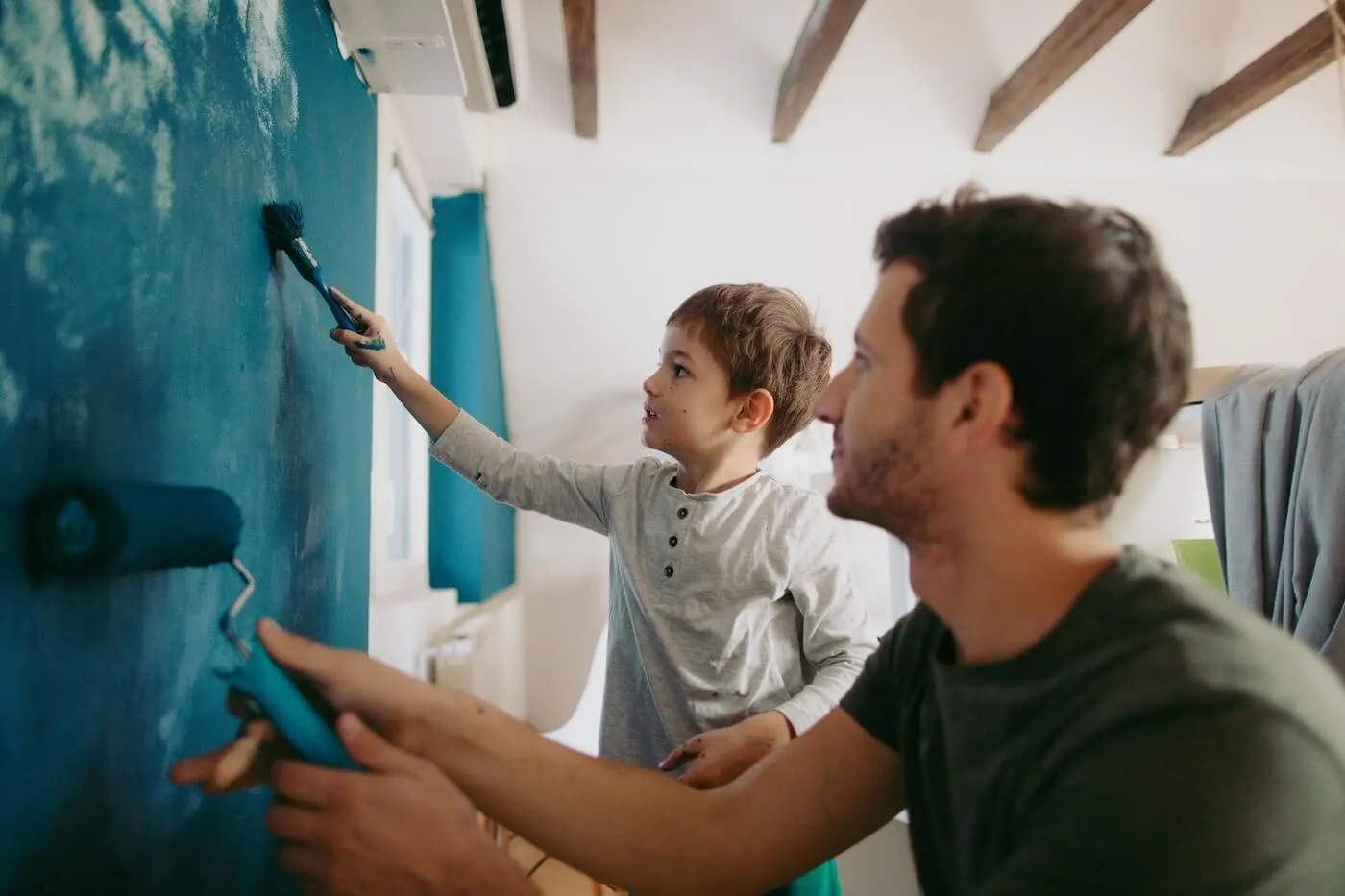 Father and toddler son painting a wall deep blue together, with paint rollers in hand.
