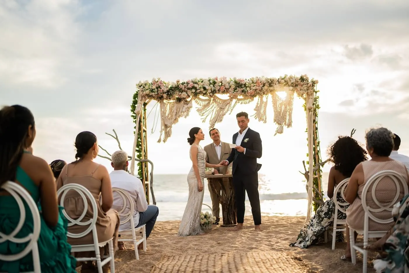 Beautiful beachside wedding ceremony at sunset, with the bride and groom standing under a flower arch exchanging vows, while guests watch and enjoy the moment