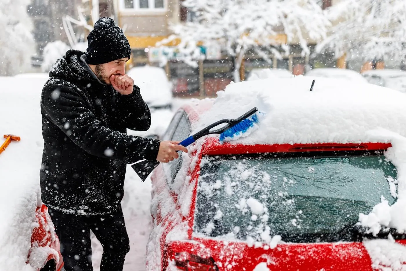 A young man feels chilly while brushing snow off his red car