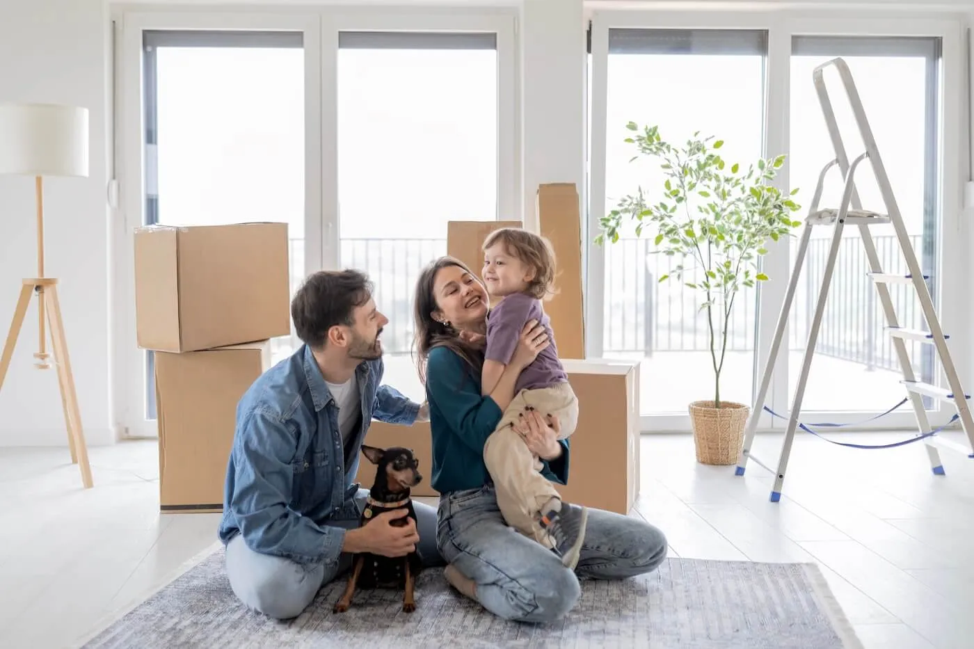 Happy family of three with a small dog sitting on the floor among moving boxes, plants, and a ladder.