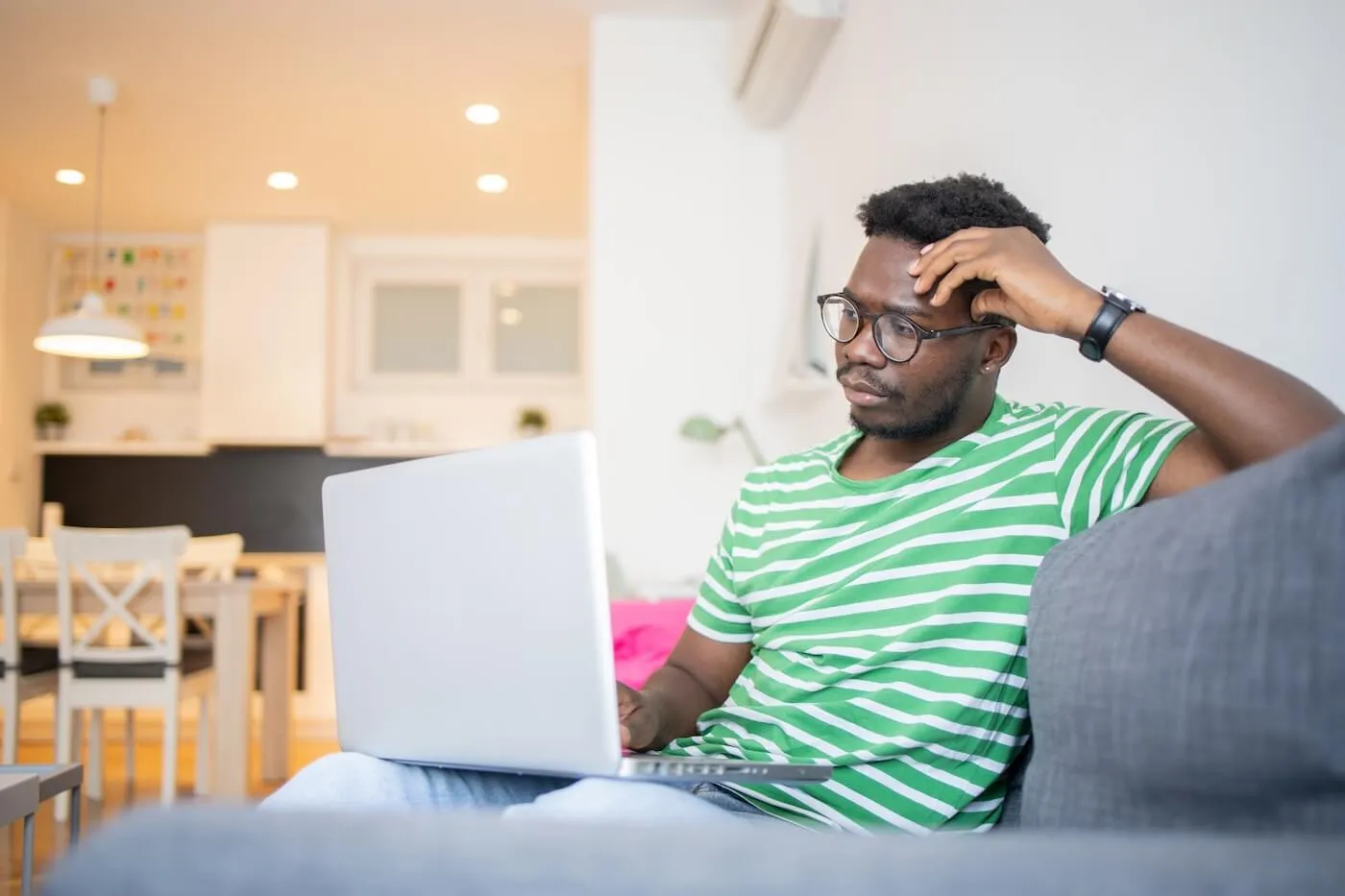 Focused man sitting on a sofa in a living room using a laptop, with a dining area visible in the background and natural light illuminating the home interior.