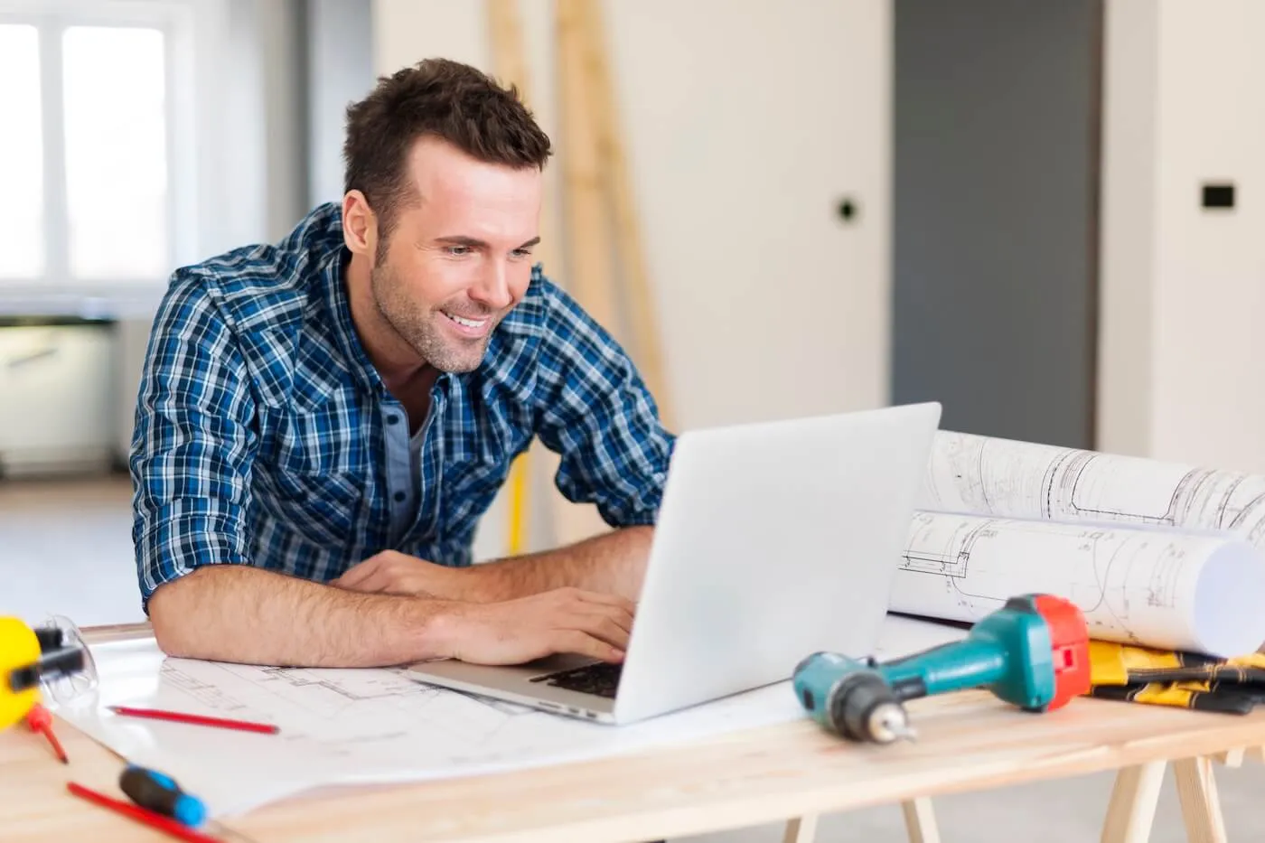 Smiling man using a laptop an a desk surrounded by tools and blueprints
