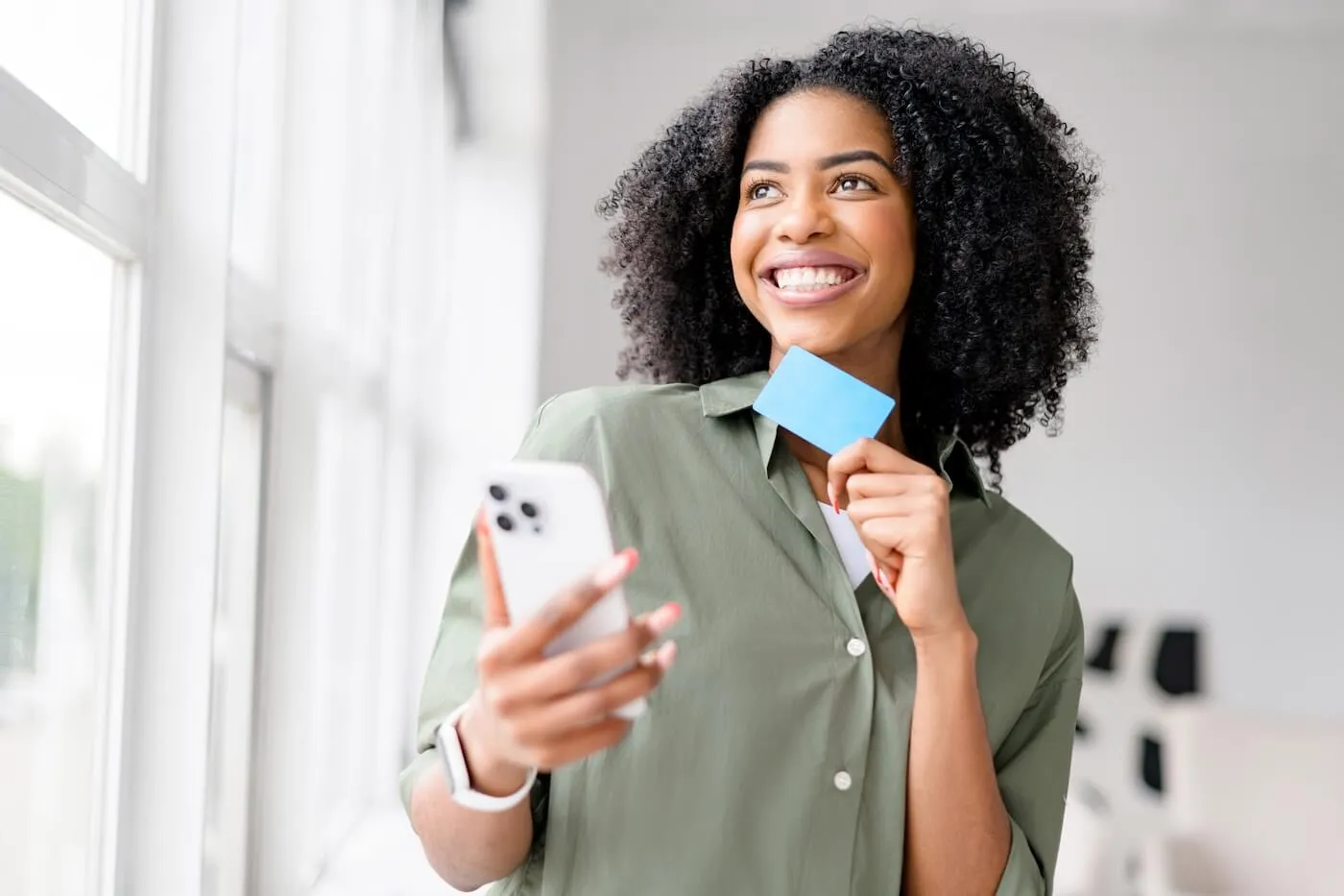 Happy young woman standing near a window in a bright home setting, holding a smartphone in one hand and a credit card in the other. The image illustrates mobile banking, online shopping, digital payments, or managing finances with a mobile device.
