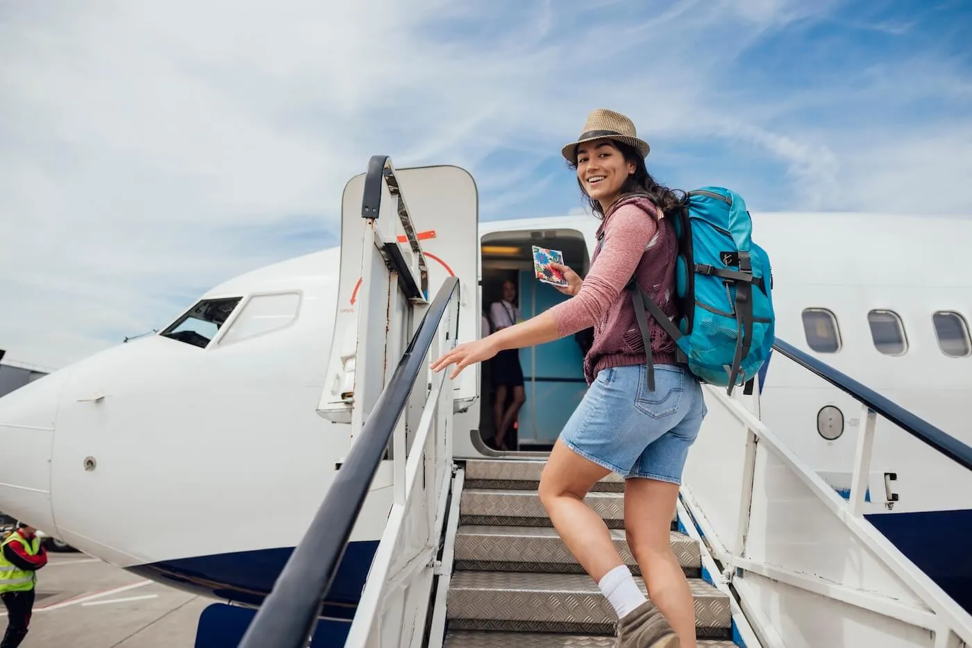 Smiling female backpacker holding a passport while boarding a plane, with a female flight attendant standing at the aircraft door