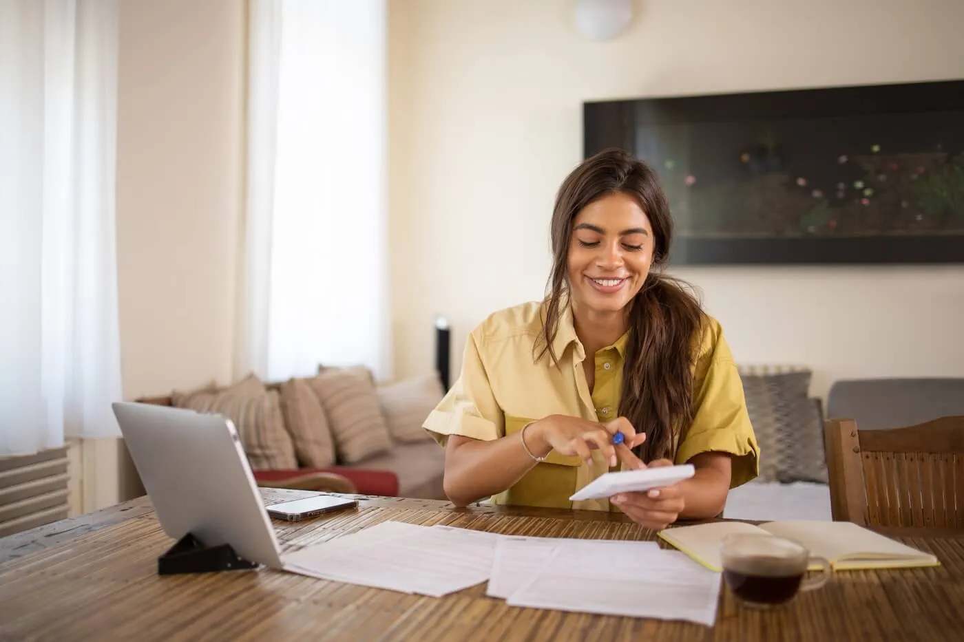 Smiling young woman budgeting at home using a calculator, tablet, and printed documents on her desk