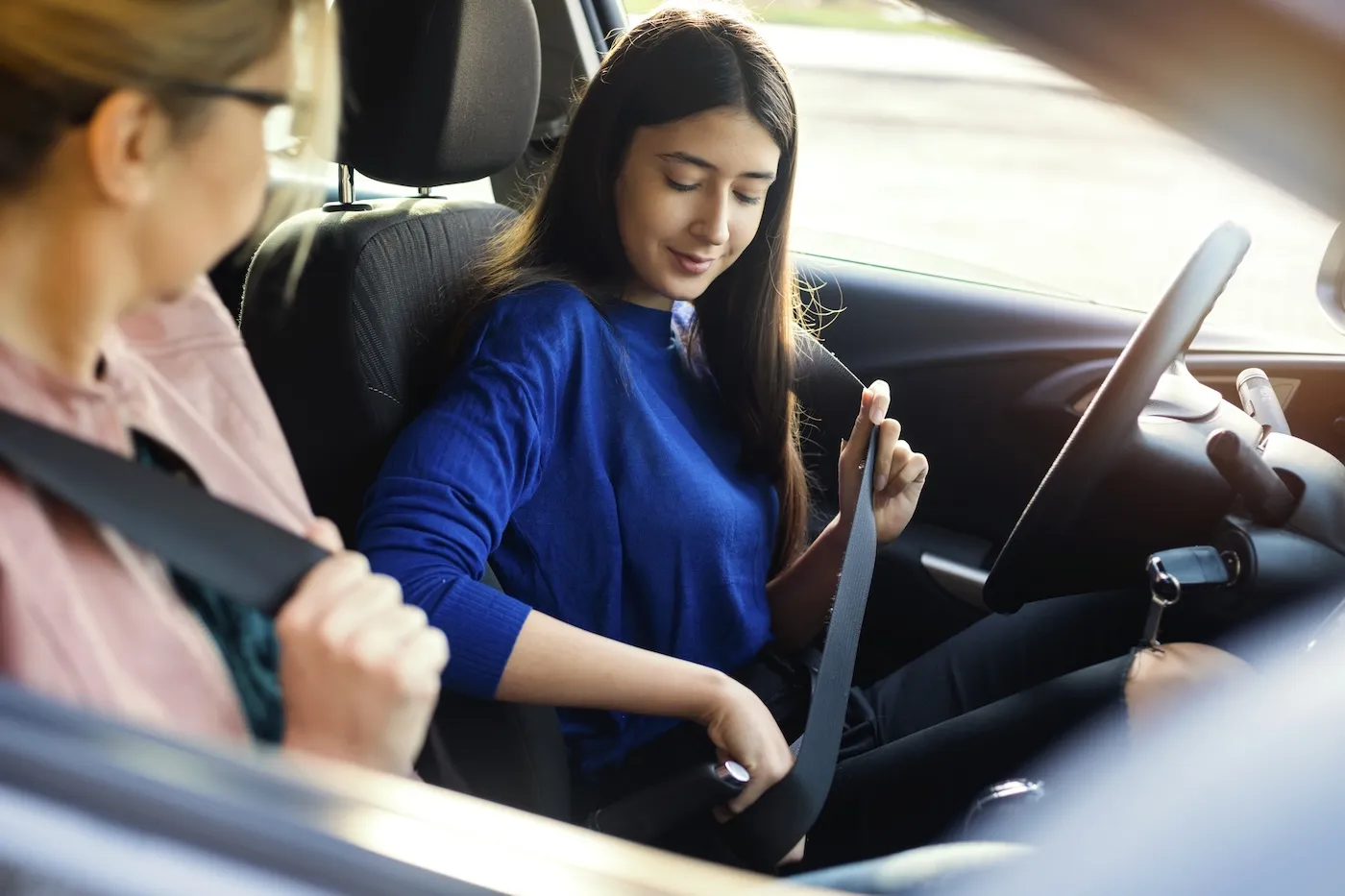 A teenage girl fastening the seatbelt on a driver's seat in the car with a mature woman on a passenger seat