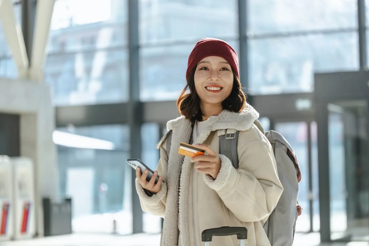Smiling woman in a winter outfit with a backpack, standing in an airport holding a smartphone and a credit card