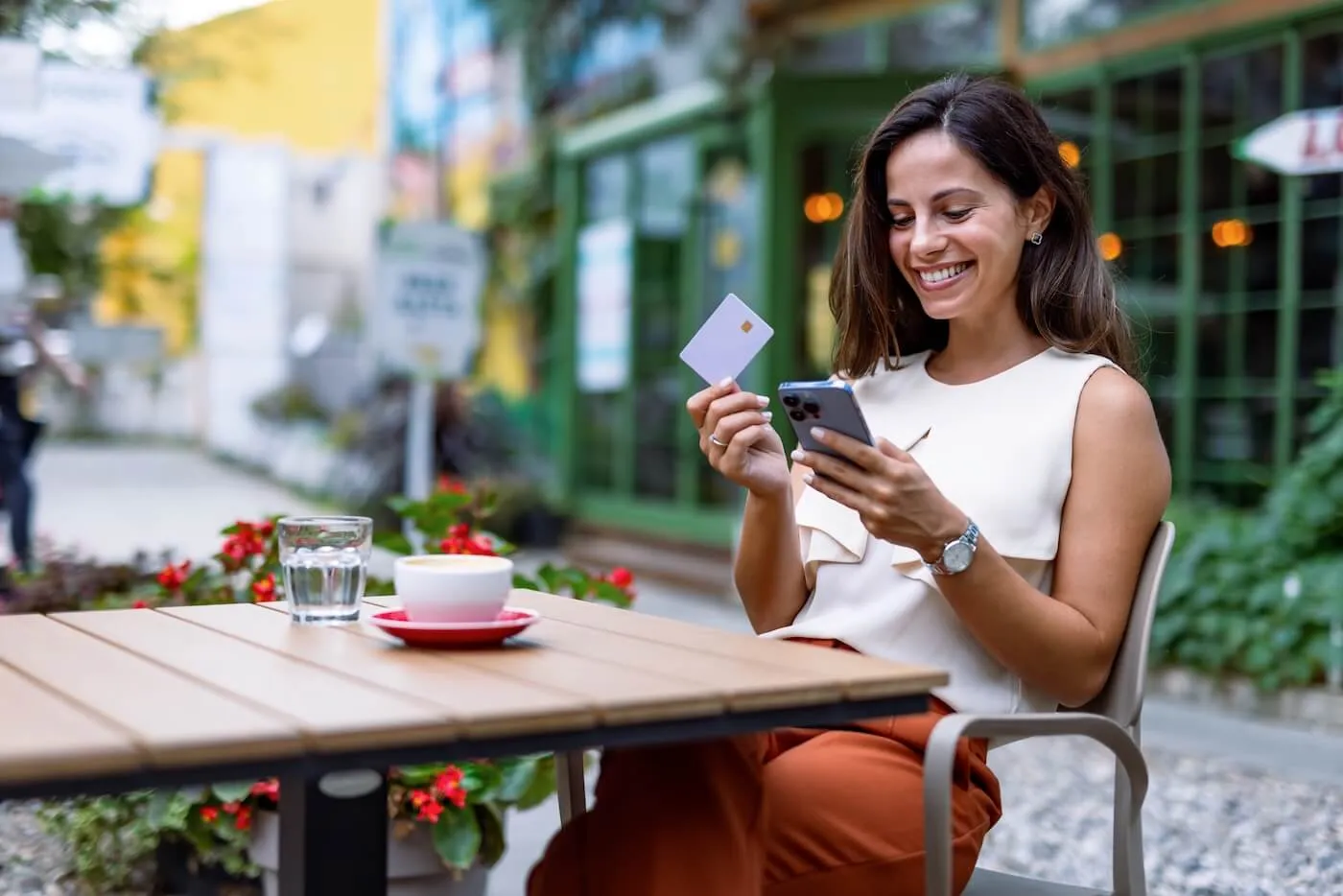 Smiling woman is using her phone and holding her credit card while enjoying coffee in the outdoor cafe