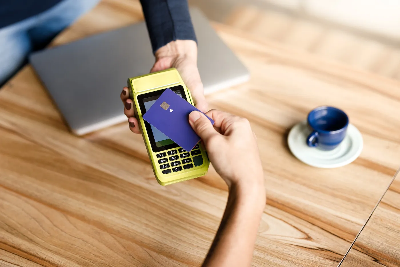 A hand holding a purple debit card up to a POS device at a cafe.