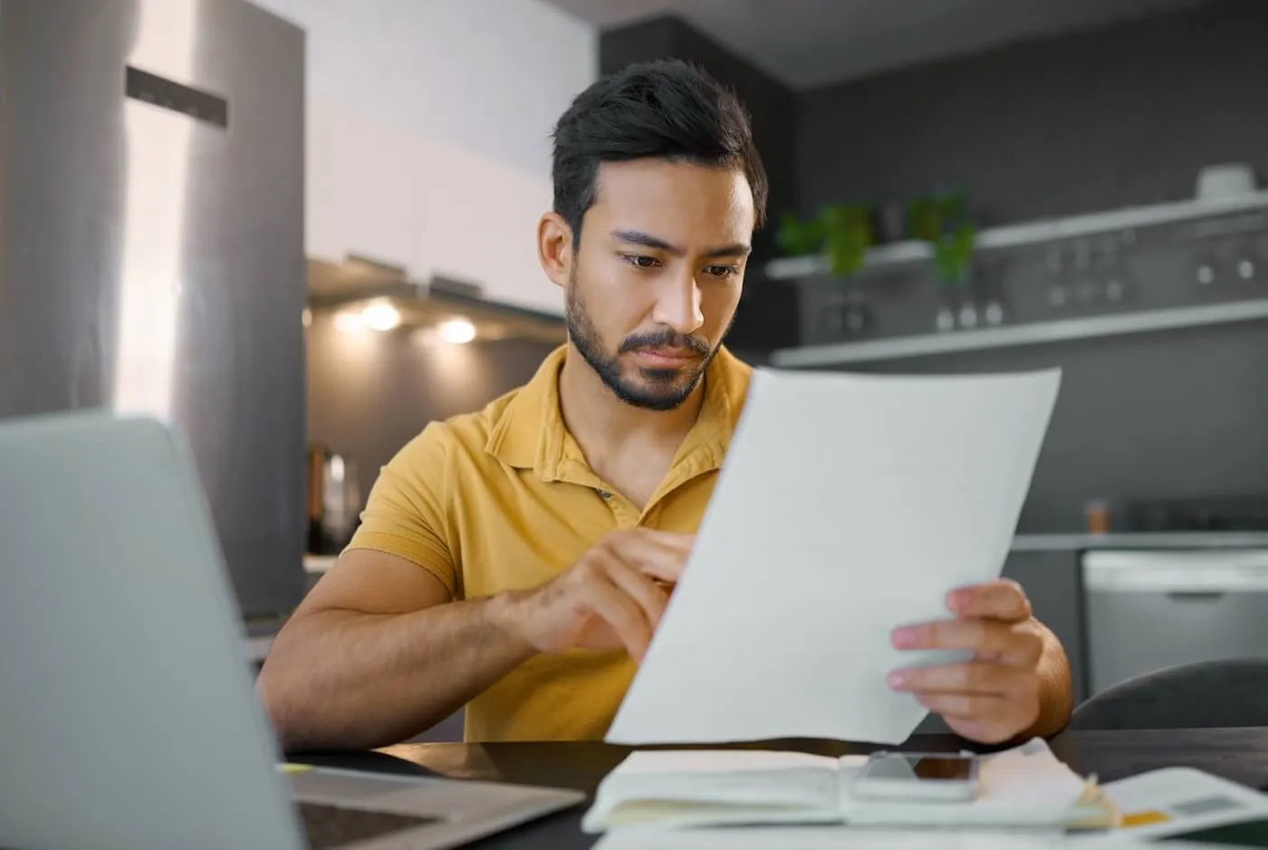 A man sits at his kitchen table reviewing debt payoff plans using a laptop and printed financial documents