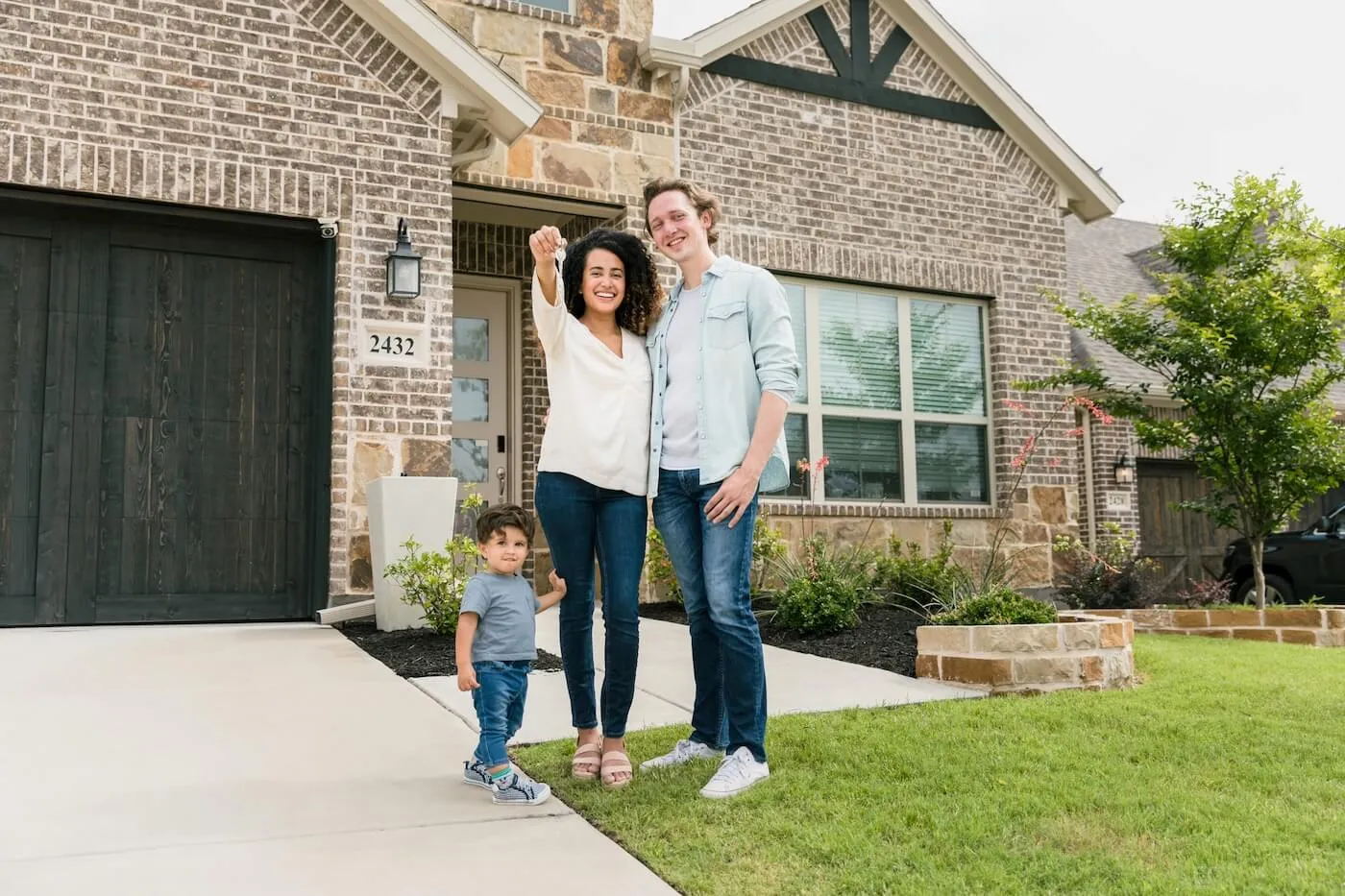 Happy family with a toddler boy standing outside of their new house and holding the keys.