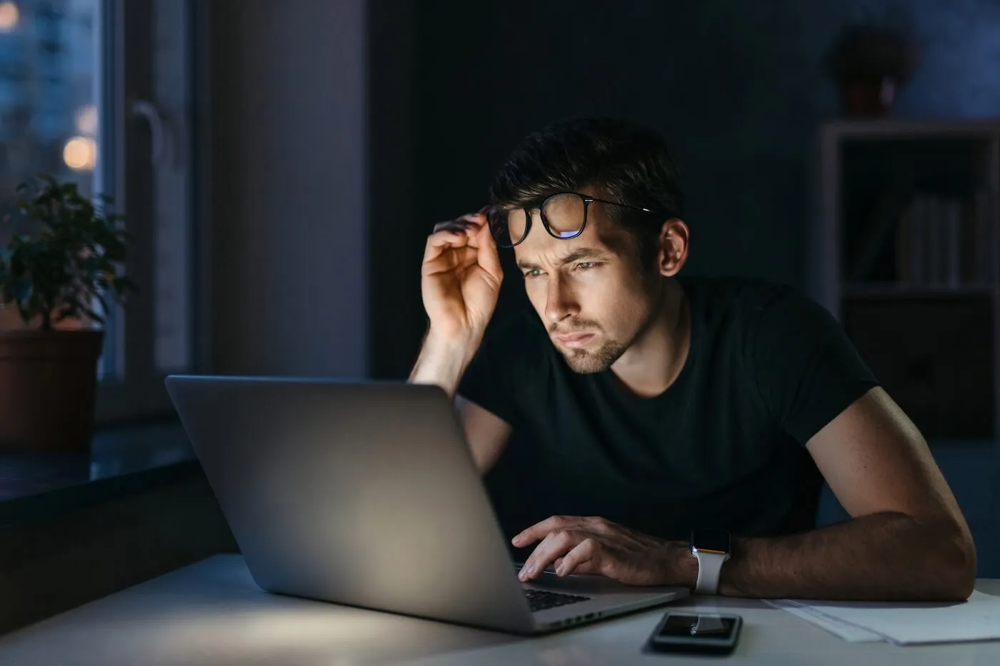 Concerned man seated at a desk in a dimly lit room uses a laptop, with one hand touching a pair of glasses and a smartphone lying nearby on the desk, while a window, shelves, and soft interior lighting are visible in the background.