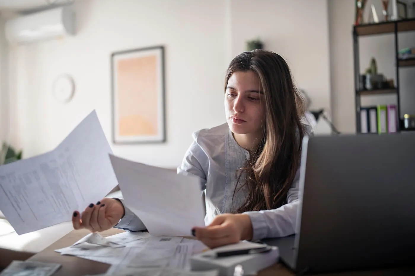 Focused young woman reviewing her bills while using a laptop at home
