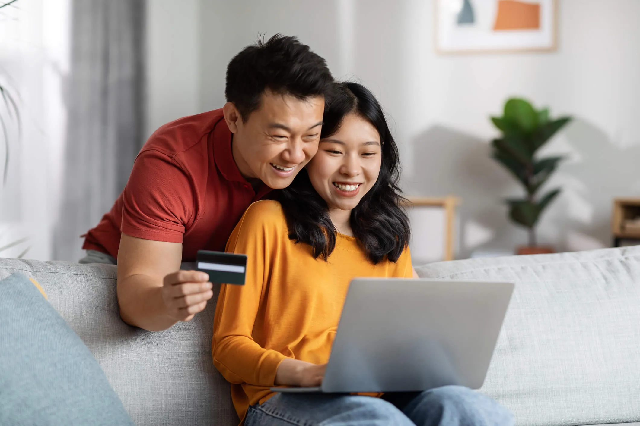 A smiling woman sits on a couch using a laptop while her partner stands close behind, holding a credit card toward the laptop.
