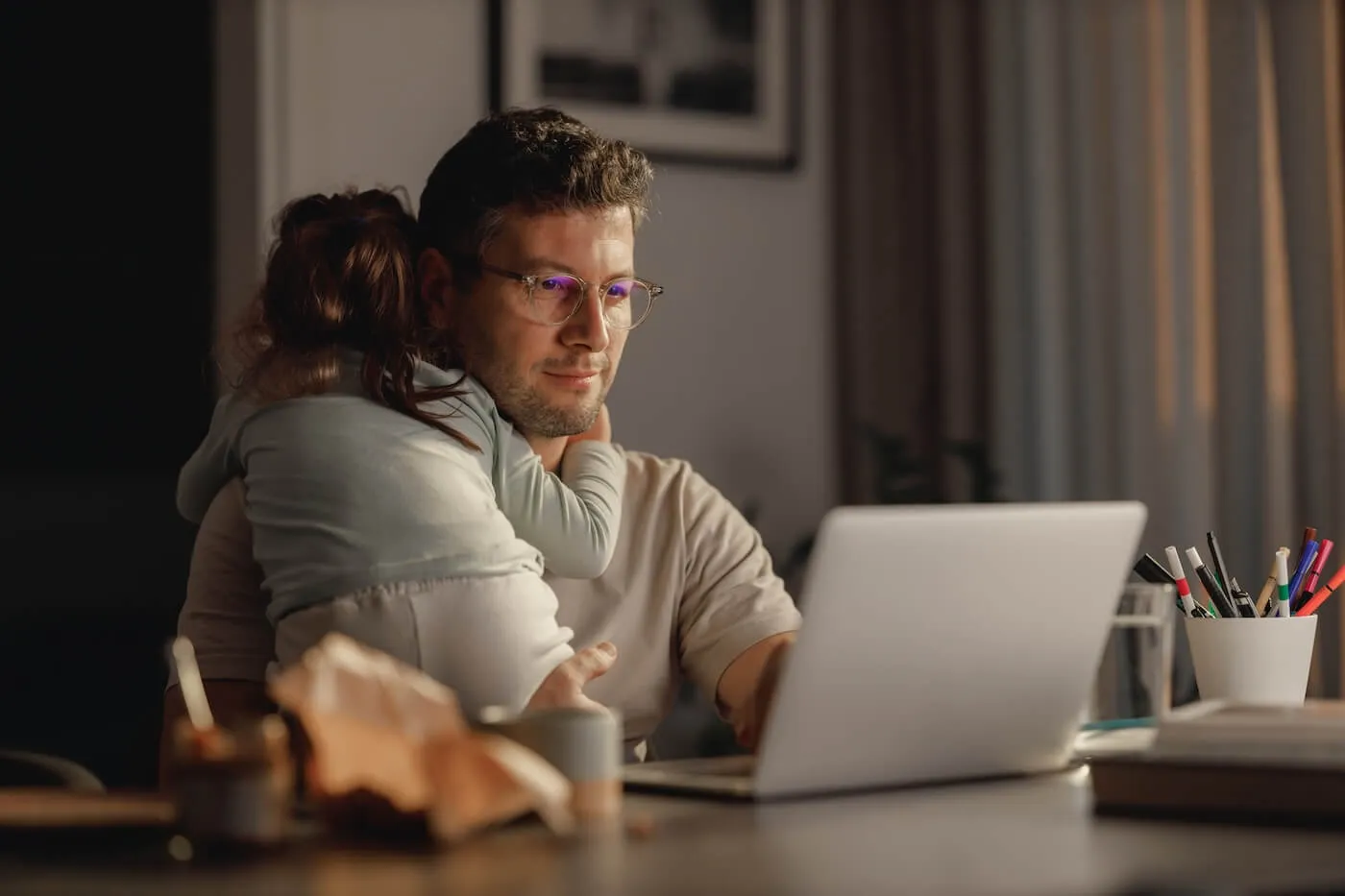 A man holding a sleeping baby daughter while using his laptop at home
