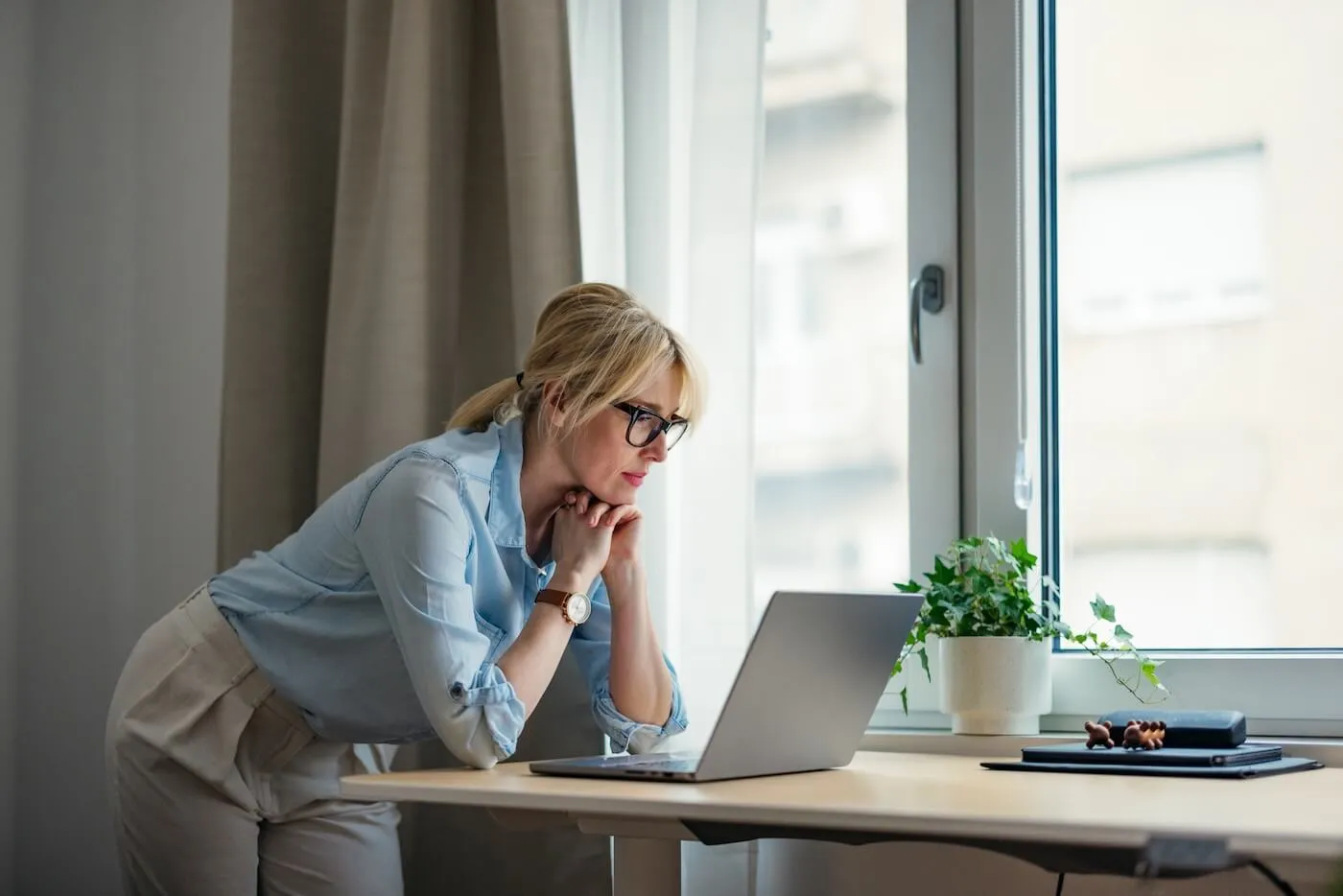 Focused woman in business attire leaning over a desk, intently viewing a laptop screen in a home office