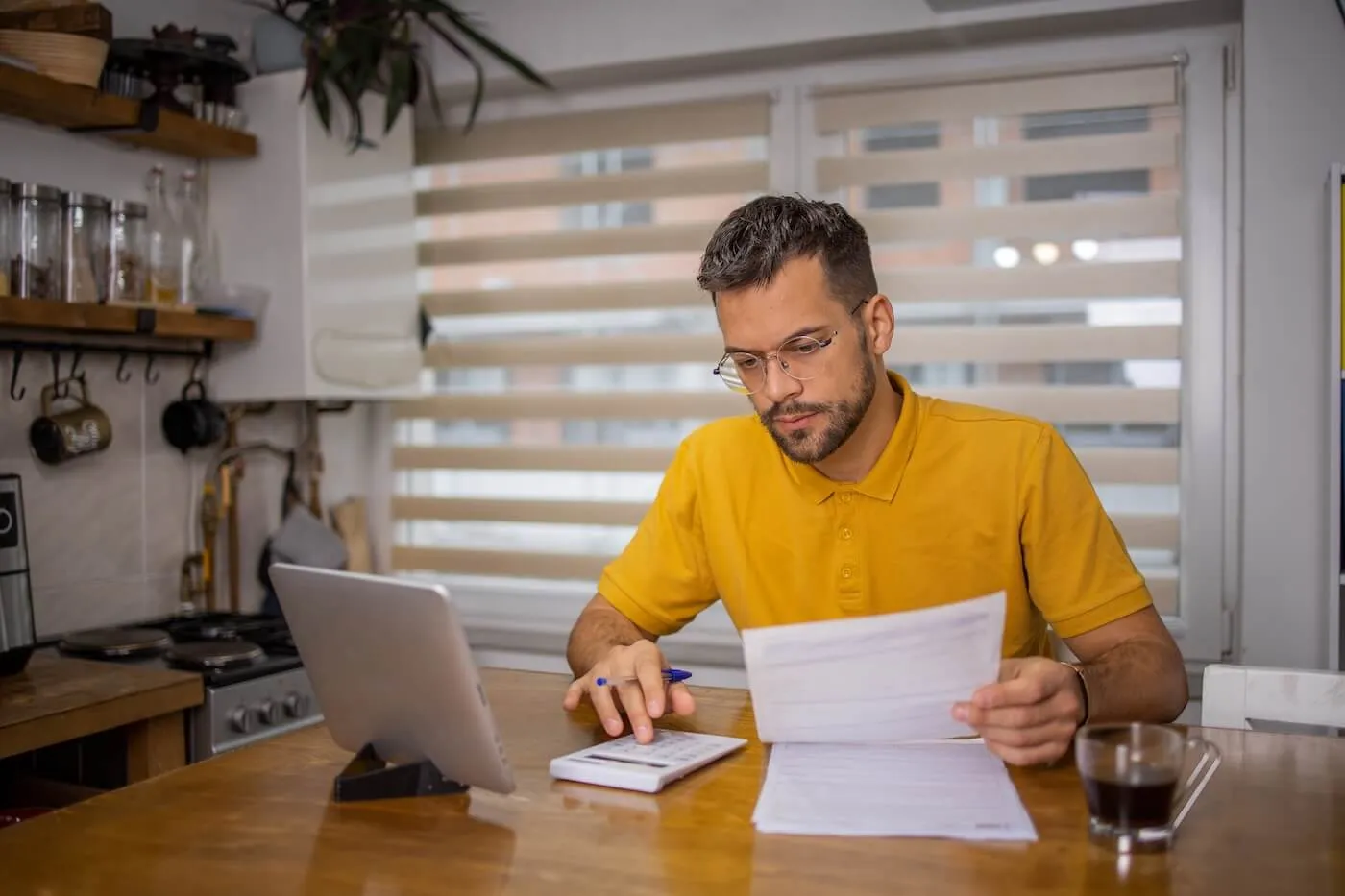 Focused man sitting at the dining table and using calculator while holding a printed document, with a tablet standing next to him