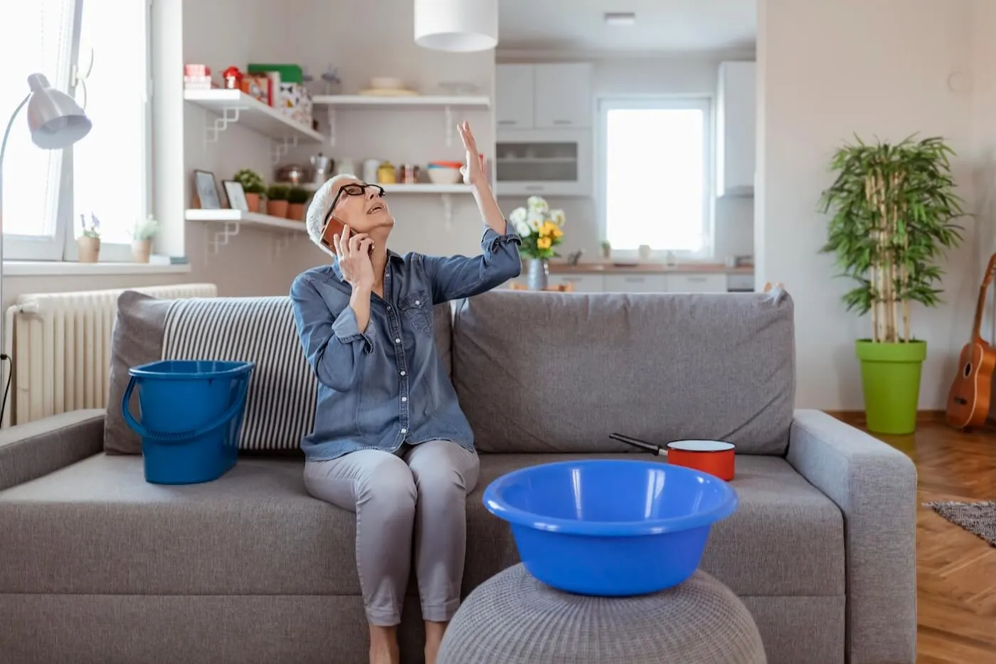 A mature woman sitting indoors, surrounded by pots and containers catching water from a leaking roof, as she talks on the phone with her insurance company