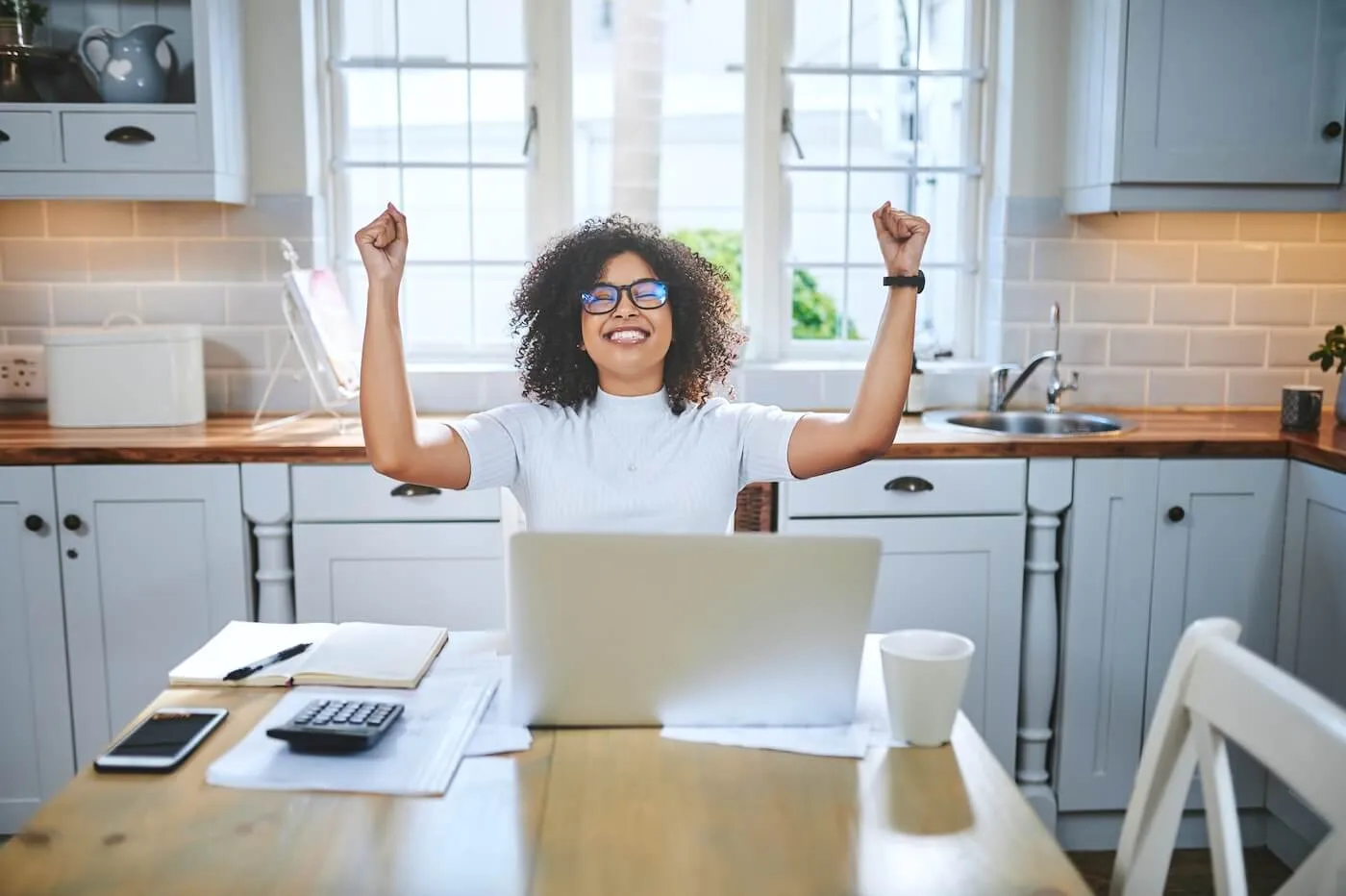 Happy young woman sits at a kitchen table with a laptop, raising both arms in a celebratory gesture, with papers, a calculator, and a phone spread out on the table