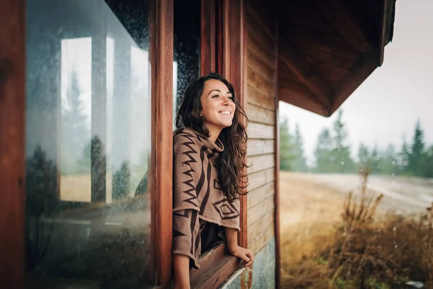 Happy young woman leaning toward the windowsill of a vacation house and observing the landscape on a rainy day