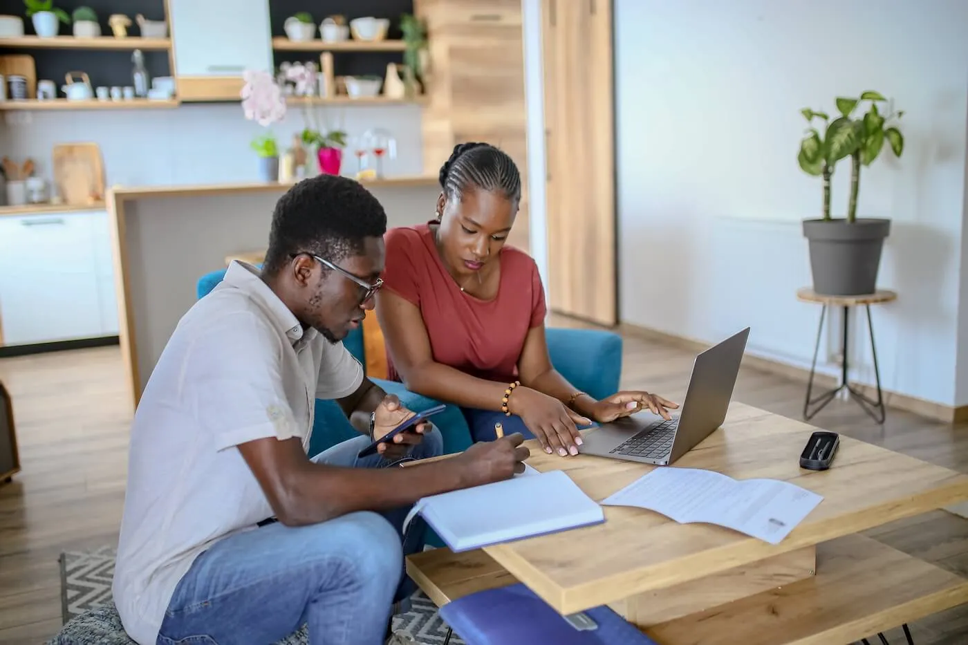 A couple sitting in their living room planning a budget— the woman is focused on a laptop while the man takes notes beside her