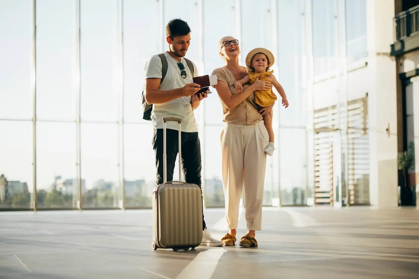 Happy family with a toddler girl standing at the sunny airport terminal; the man with a suitcase checking the tickets while the woman is holding a child