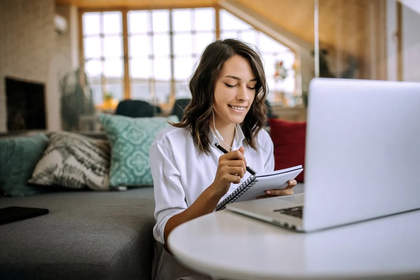 Smiling young woman in headphones using her laptop and making notes at home