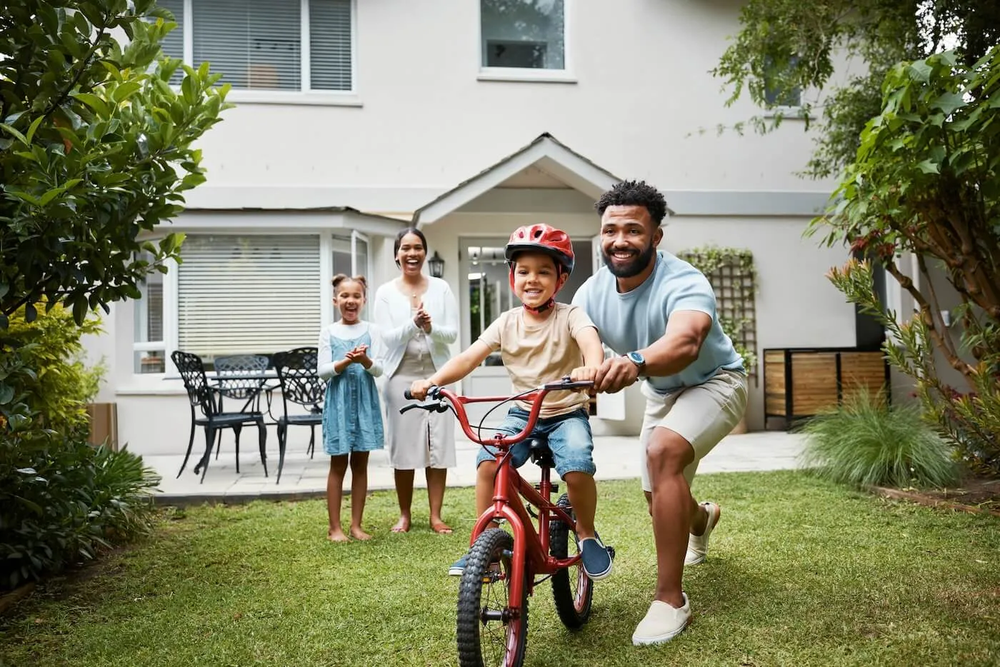 Happy family of four enjoying time in the green backyard, with a mother and daughter applauding a young boy riding a bicycle while his father supports him
