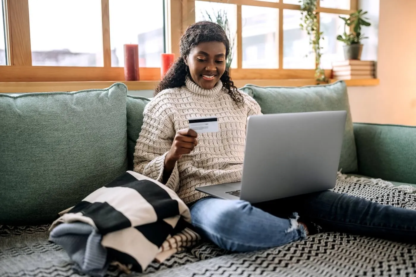 Smiling young woman sitting on a sofa with a credit card and laptop, cozy plaid blanket beside her.