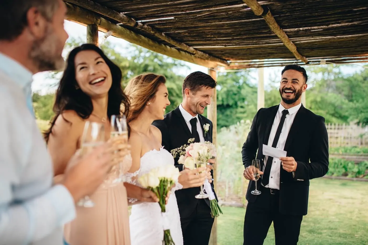 Group at an outdoor wedding reception holds flowers and drinks while one person gives a speech under a wooden canopy.