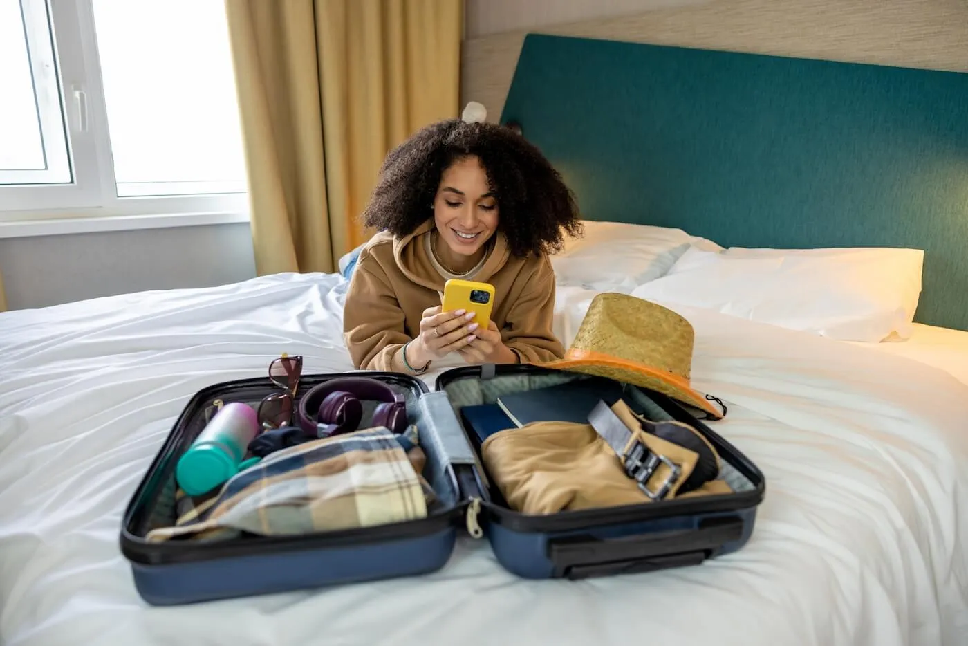 Smiling young woman lying on a bed with a packed suitcase beside her, using her smartphone to buy last-minute travel insurance