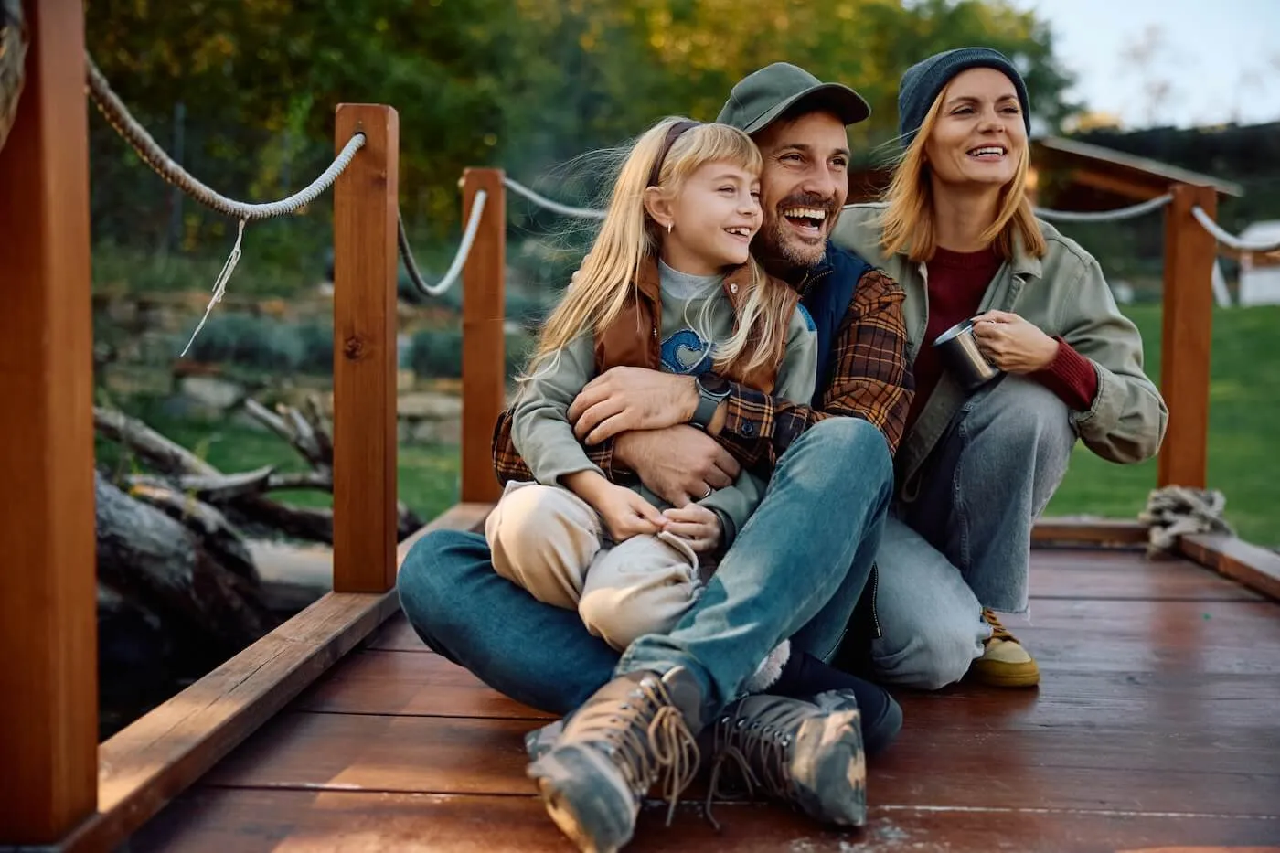 Happy family of three sitting on the outdoor wooden deck, the woman is holding a travel mug