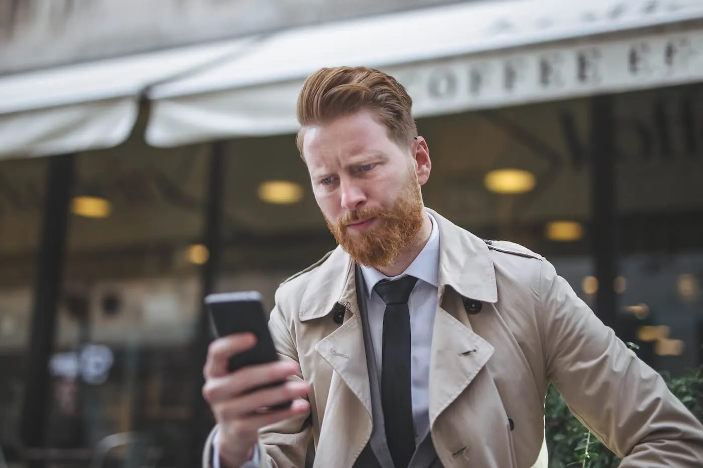 Concerned man seated at an outdoor café looking at a smartphone while wearing a trench coat and business attire, with a coffee shop storefront blurred in the background.