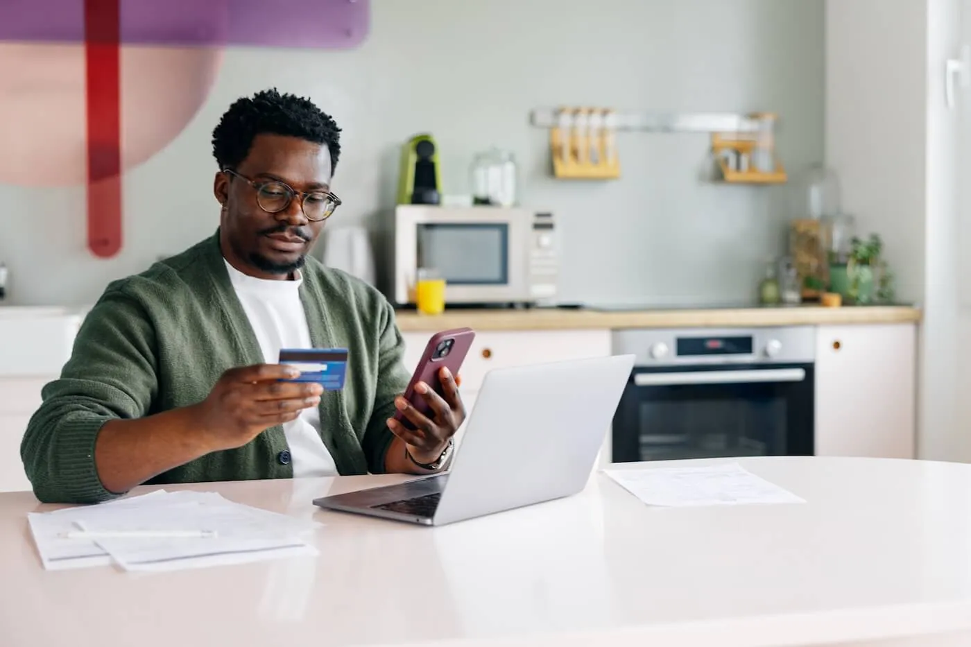 Focused man seated at a kitchen table holding a credit card and using a smartphone beside an open laptop. Documents are spread across the table, with kitchen appliances, countertops, and cabinets visible in the background.