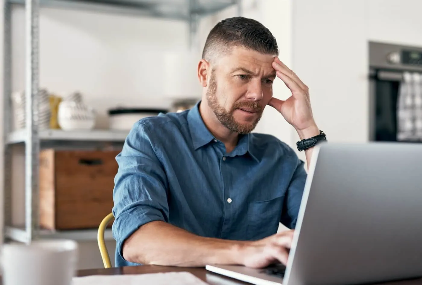 Concerned man sitting at a table in a home setting, working on a laptop with one hand on the keyboard and the other resting on their forehead, with shelves and household items visible in the background.