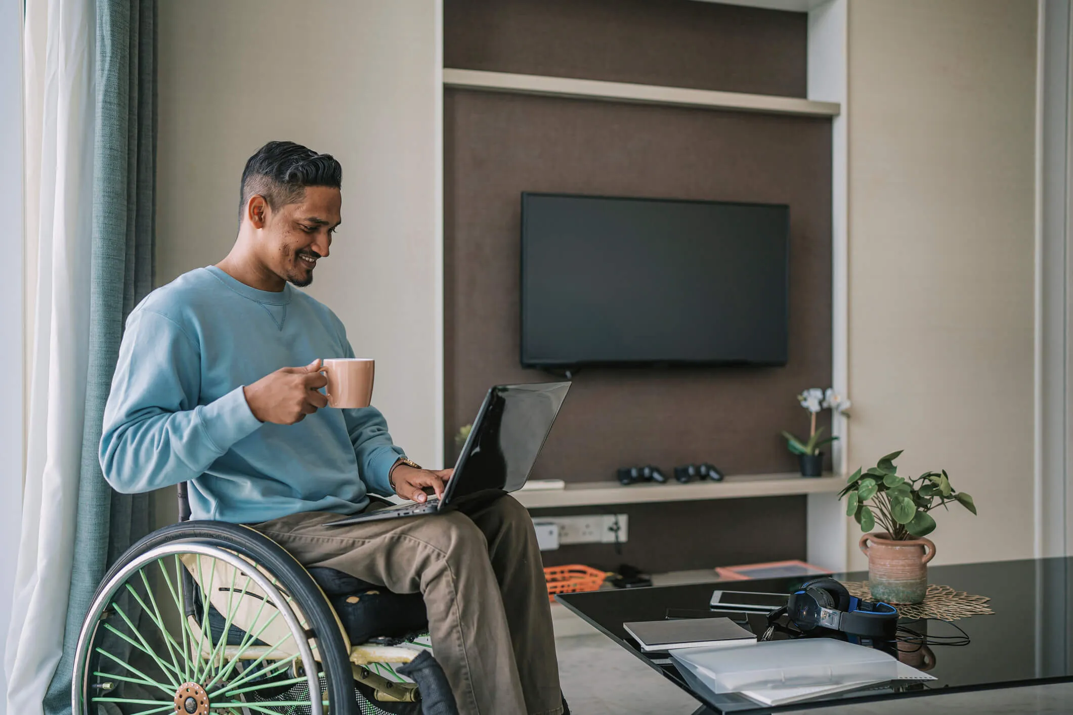 Smiling young man on a wheelchair using his laptop at home while enjoying a hot drink