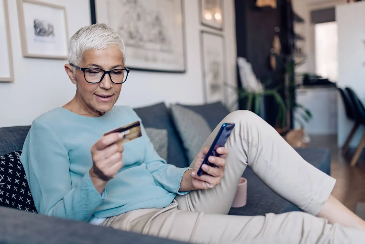 Senior woman sits on a couch holding a credit card and using a smartphone, with framed artwork and home furnishings in the background.
