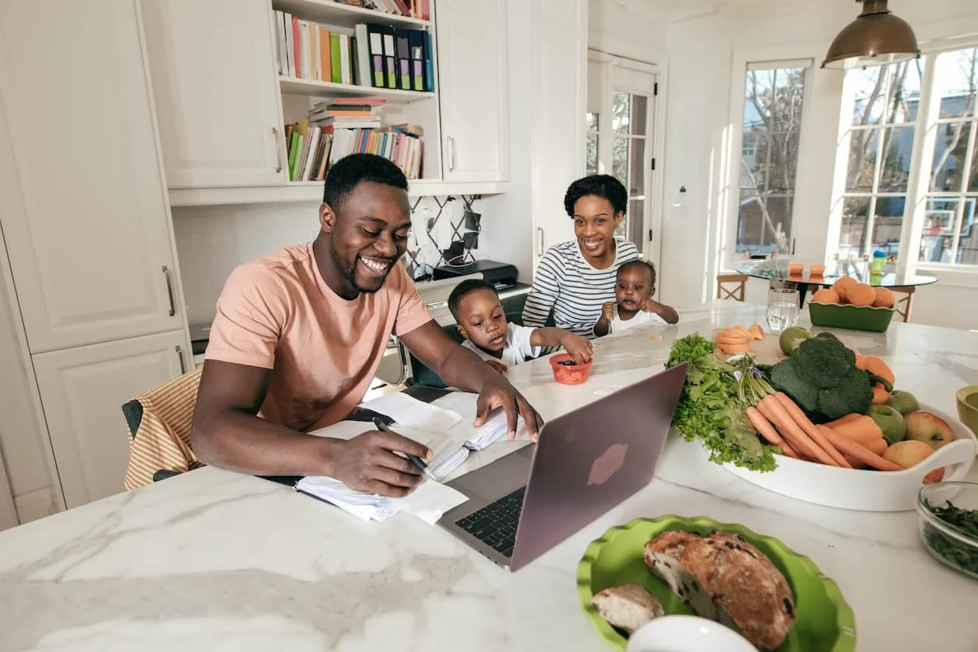 Happy family with two young kids are filing taxed online in the kitchen, plates with fruit and vegetables standing on the table beside them