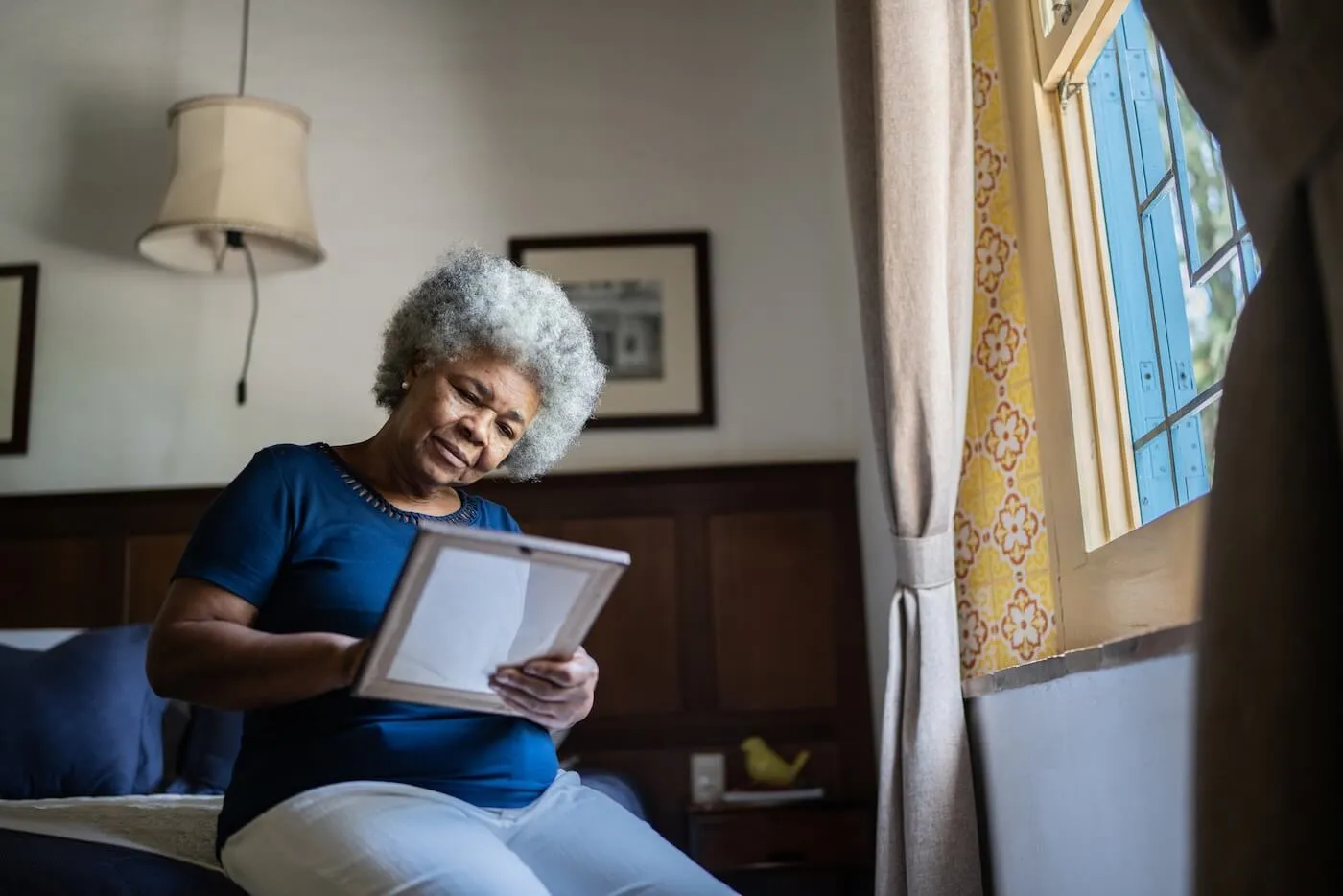 Older woman seated on the edge of a bed near a window, holding and looking at a framed photograph. The bedroom features wood-paneled furniture, framed artwork, patterned wall accents, and natural light streaming through an open window