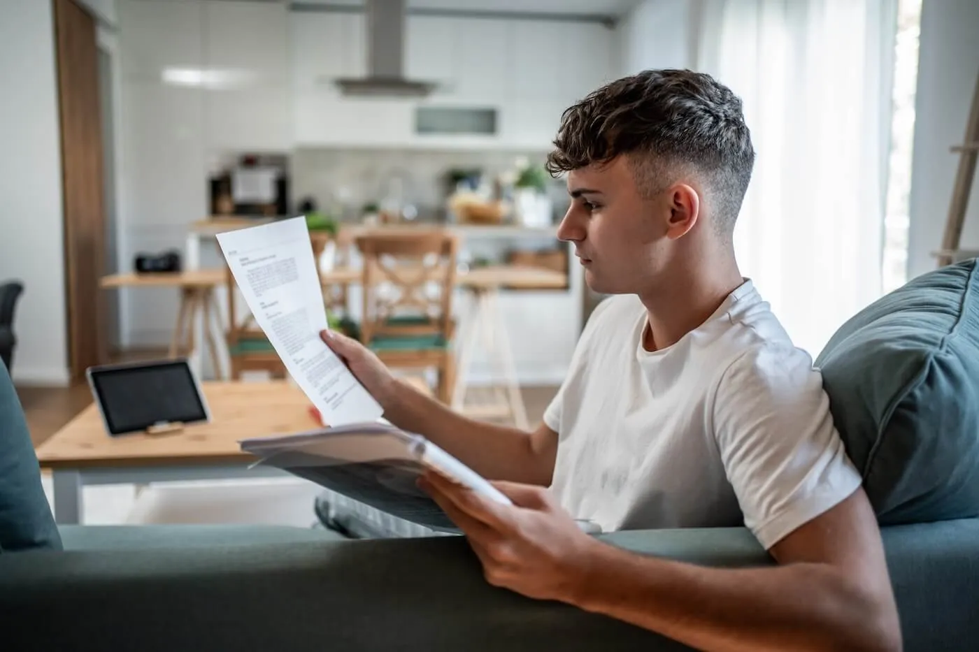 Focused young man sitting on a sofa and reviewing printed documents; a tablet and kitchen area are visible in the background.