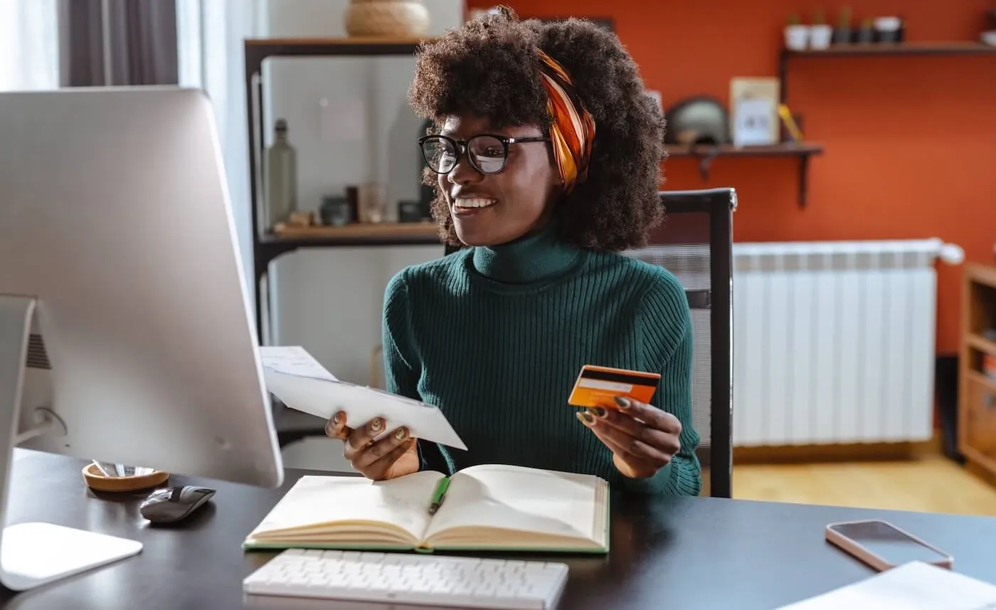 Young woman sitting at her home desk, holding a credit card and a letter, looking at her computer monitor with a cheerful expression
