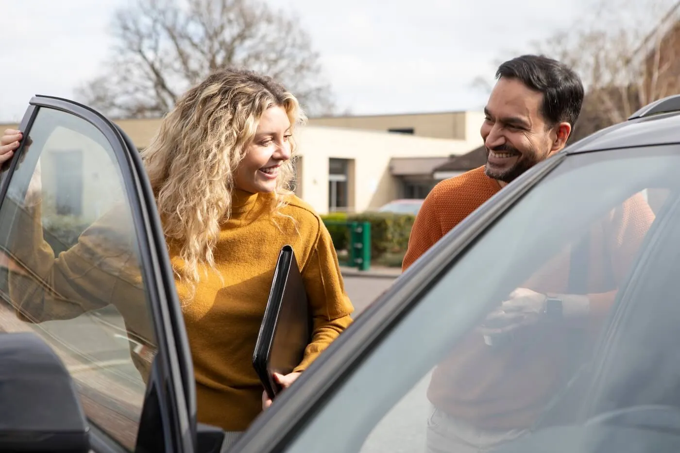A smiling man and a woman are opening a car door, the woman is holding a folder