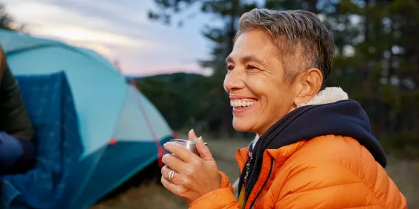 Person holding a metal mug near a tent outdoors at dusk, wearing an orange jacket with trees in the background.